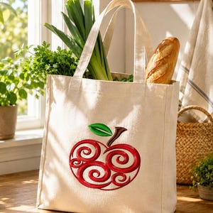 May include: A cream-colored canvas tote bag featuring an embroidered red apple design with swirling patterns and a green leaf. The bag is filled with fresh green leeks and a crusty baguette, showcasing a structured rectangular shape with two handles.