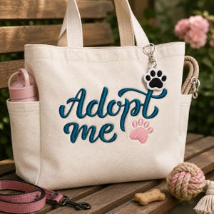 May include: A cream-colored canvas tote bag featuring the embroidered text "Adopt me" in dark teal script, accompanied by a pink embroidered paw print. A white and black paw print keychain hangs from the handle, and a pink water bottle is in the side pocket.