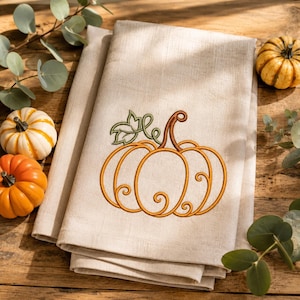 Può includere: Un tovagliolo di lino beige piegato con una zucca arancione ricamata, dotata di un picciolo marrone e foglie verdi. Il disegno della zucca è delineato da volute decorative. Il tovagliolo è presentato con piccole zucche decorative ed eucalipto.