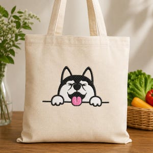 May include: A natural-coloured canvas tote bag featuring an embroidered graphic of a black and white husky dog peeking over a horizontal line with its pink tongue sticking out and eyes closed in a happy expression, complete with two shoulder straps.