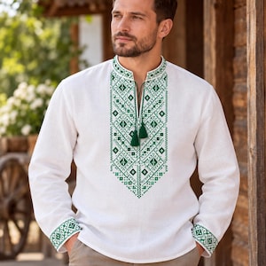 Pode incluir: Uma camisa branca de manga comprida com um padrão geométrico bordado a verde no centro do peito e nos punhos. A camisa tem uma gola mandarim com decote aberto e duas borlas verdes penduradas em cordões finos verdes no pescoço.