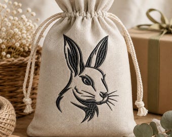 Può includere: Un sacchetto con coulisse beige in tessuto intrecciato con un ricamo nero raffigurante una testa di coniglio sul davanti. La borsa è dotata di due spesse corde intrecciate color crema che si tirano per chiudere la parte superiore arricciata.