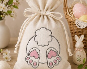 Può includere: Un sacchetto color crema con chiusura a coulisse in tessuto intrecciato, caratterizzato da un ricamo che raffigura una coda di coniglio e due zampe con cuscinetti rosa a forma di cuore. L'apertura è arricciata da un cordino e rifinita con pizzo.