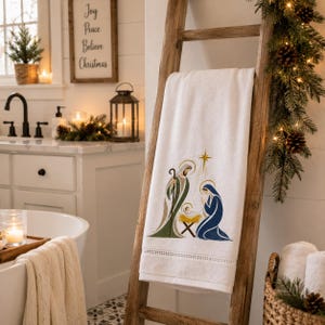 May include: A white hand towel featuring an embroidered nativity scene with figures in green, blue, and gold thread. The design depicts a stylized holy family with a star above, finished with a decorative hem detail at the bottom edge.