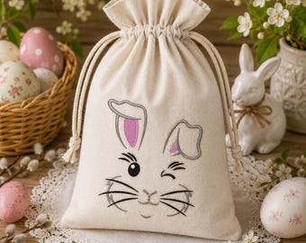 Può includere: Un sacchetto con coulisse in tela beige caratterizzato da un ricamo raffigurante il muso di un coniglio che fa l'occhiolino, con orecchie interne rosa, occhi neri e baffi. La borsa presenta una chiusura arricciata con due spessi cordini beige.