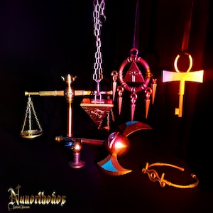 May include: A collection of golden Egyptian-themed jewellery and accessories, including a scale, an axe, a crown, a pendant with an eye, and a key. The items are arranged on a black background.