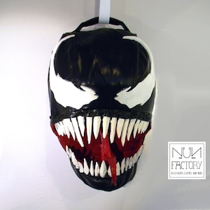 May include: A black and white Venom mask with a red tongue hanging out. The mask is made of a soft, flexible material and has a detailed design. The mask is hanging from a hook on a white wall. The text "NUM FACTORY" is visible in the bottom right corner of the image.