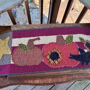 Puede incluir: Un camino de mesa acolchado rectangular con un fondo de rayas color burdeos y crema. Los elementos aplicados incluyen una estrella amarilla, dos calabazas de cuadros marrones y rojos, dos girasoles con centros oscuros y una silueta de cuervo negro.