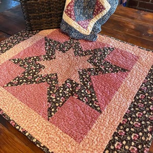 May include: A handmade quilt featuring a star design in pink, black, and cream hues. The quilt is displayed on a wooden surface, with a basket and other quilted items in the background. The star pattern is the main feature.