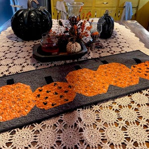 May include: A rectangular quilted table runner featuring four orange pumpkin-shaped fabric appliqués with black patterns on a dark gray textured background. Each pumpkin has a small black stem at the top, and the entire runner has a black border.