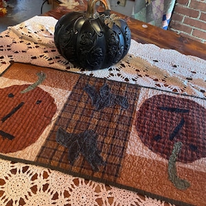 May include: A rectangular quilted table runner featuring three distinct panels. The outer panels display orange and dark red pumpkin shapes with stitched details, while the center panel features a brown plaid fabric with two dark bat silhouettes.