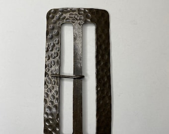 May include: A rectangular metal belt buckle with a dark, hammered finish. The buckle features a central vertical bar and a thin, hinged prong that extends across the left opening. The surface has a textured, dimpled appearance throughout.