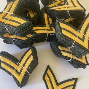 May include: Stacks of black and gold chevron patches, each with two gold stripes. The patches are bundled together with thin, clear elastic bands, suggesting they are ready for use or sale. The patches are likely for uniforms or military-themed projects.