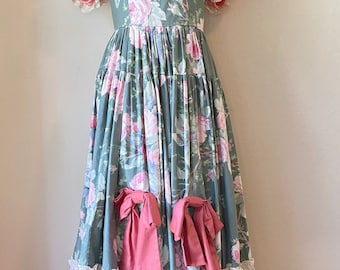 May include: A floor-length dress featuring a sage green fabric with a pink floral pattern, short ruffled sleeves with lace trim, a square neckline, and a matching choker. The skirt includes two pink fabric bows and a wide solid pink bottom tier.