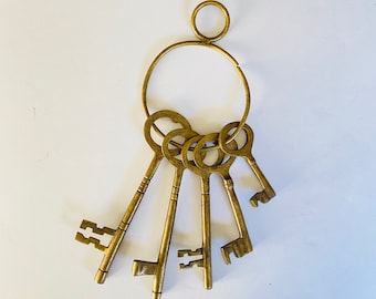 May include: A set of antique-style brass keys on a large ring. The keys have intricate designs and varying shapes, with a circular ring at the top for hanging. The keys are arranged in a fan shape, suggesting a decorative or collectible item.