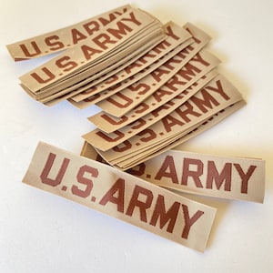 May include: A stack of tan fabric patches with the text "U.S. ARMY" printed in brown. The patches are rectangular and appear to be new. The image is taken from a top-down perspective.