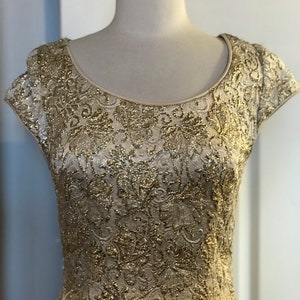 May include: A gold-colored, short-sleeved dress with an intricate floral pattern. The dress is displayed on a mannequin, showcasing its scoop neckline and textured fabric. The dress appears to be made of a shimmering, possibly metallic material.