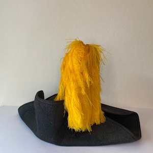 May include: A dark gray tricorn hat with a bright yellow feather plume. The hat has a wide brim and a classic, historical design. The feathers are clustered and add a striking contrast to the hat's color.