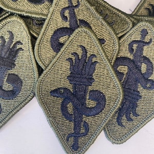 May include: A collection of olive green diamond-shaped embroidered patches. Each patch features a dark blue emblem of a serpent coiled around a staff, with a sword. The patches are densely packed, showcasing the detailed stitching and design.