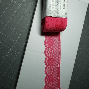 May include: Pink lace trim with a floral pattern. The trim is packaged on a roll with a label that reads "Color 248", "Lot 002", and "3.21.00".