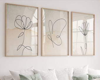May include: Three minimalist line art prints of flowers in black on a white background with a watercolor wash in beige and light green. The prints are framed in natural wood and hung on a white wall above a white couch with green and beige throw pillows.