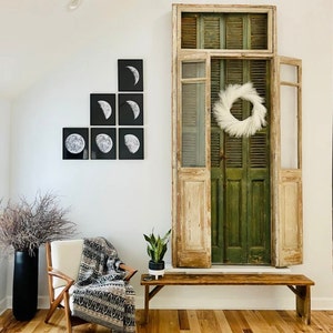 May include: A white wall with a set of black and white framed prints of moon phases. A wooden bench sits in front of a large, weathered, green wooden door with a white wreath hanging on it. A potted plant sits on a small table next to the bench.