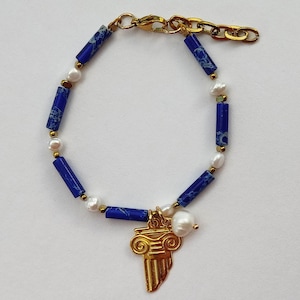 Puede incluir: Una pulsera dorada con cuentas cilíndricas azules y blancas, pequeñas perlas y un colgante dorado con forma de columna clásica. La pulsera está conectada por una cadena y un cierre dorados.