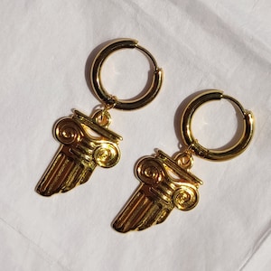 May include: Gold hoop earrings with a gold winged column charm hanging from each hoop.