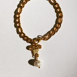 Puede incluir: Un brazalete de cadena dorada con un dije dorado en forma de columna con una perla colgando.