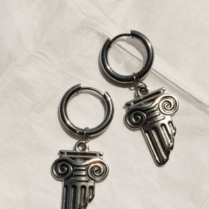 May include: A pair of silver hoop earrings with a decorative column charm. The charms feature a detailed design with scrolls and lines.