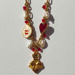 Könnte beinhalten: Eine goldene Kettenkette mit roten und weißen Perlen, einem roten Herz-Anhänger und zwei goldenen Herz-Anhängern. Einer der goldenen Herz-Anhänger hat eine weiße Emaille-Mitte mit einem roten Herzen und Dornen.