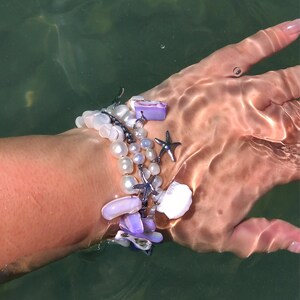 以下が含まれることがあります： 白い真珠、紫と白の貝殻のチャーム、銀のヒトデのチャームが付いたブレスレット。ブレスレットは水の中で手首につけられています。