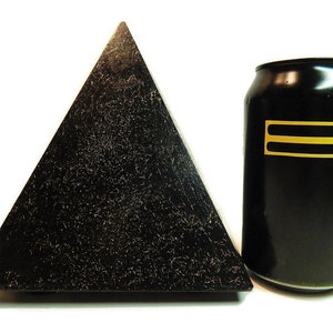 May include: A black pyramid with a textured, speckled surface next to a black aluminum beverage can. The can features two horizontal yellow stripes on its side. The pyramid has a wide base and tapers to a point, contrasting with the cylindrical shape of the can.