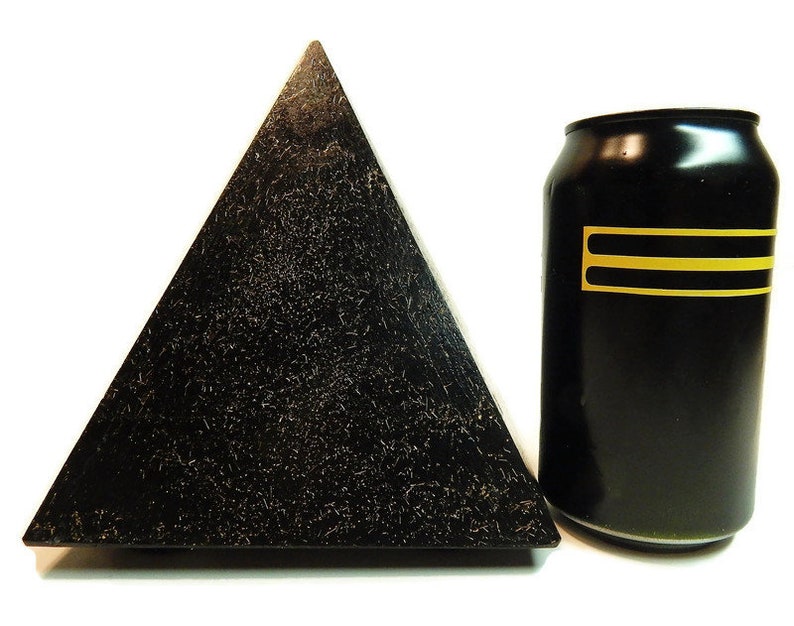 May include: A black pyramid with a textured, speckled surface next to a black aluminum beverage can. The can features two horizontal yellow stripes on its side. The pyramid has a wide base and tapers to a point, contrasting with the cylindrical shape of the can.