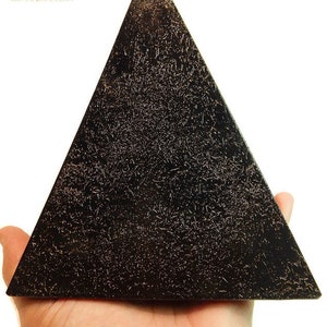 May include: A black pyramid-shaped object with a textured surface featuring fine, light-colored metallic-like flecks throughout. The top left corner displays the text KIND ORGONE with a small yellow pyramid icon and blue crystal graphics.