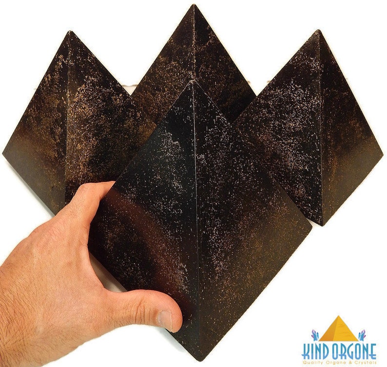 May include: Three dark brown, pyramid-shaped objects with a textured, speckled surface finish. The pyramids have a smooth, geometric form with four triangular sides meeting at a sharp apex. A logo in the corner reads Kind Orgone Quality Orgone & Crystals.