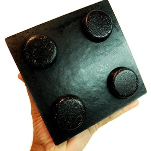 May include: A square black object with a smooth, flat surface featuring four raised, circular, textured protrusions arranged in a grid pattern. The item has a matte finish and a solid, uniform dark color throughout its entire structure.