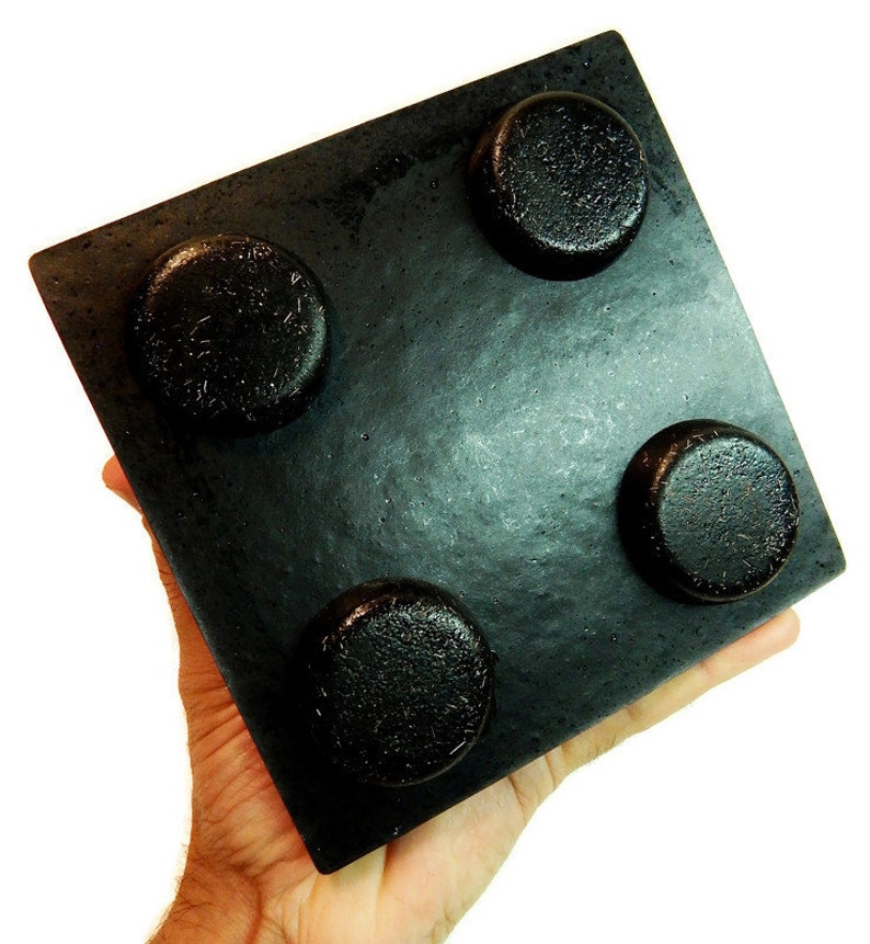 May include: A square black object with a smooth, flat surface featuring four raised, circular, textured protrusions arranged in a grid pattern. The item has a matte finish and a solid, uniform dark color throughout its entire structure.