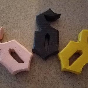 May include: Three wooden 3D printed letters, each in a different color: pink, black, and yellow. The letters are all the same design, a stylized letter 'S'.