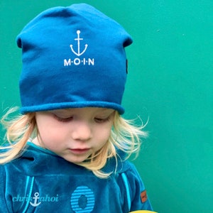 Könnte beinhalten: Blauer Beanie mit weißem Anker-Design und dem Wort "M.O.I.N" auf der Vorderseite. Der Beanie wird von einem Kind mit blonden Haaren getragen. Das Kind trägt einen blauen Kapuzenpullover. Der Hintergrund ist einfarbig grün.