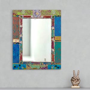 May include: Rectangular mirror with a mosaic frame in a patchwork design. The frame features blue, green, red, and yellow hues. Floral and beaded embellishments decorate the edges. A decorative wall mirror for the home.