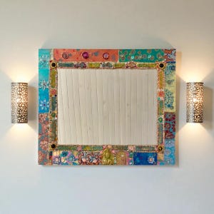 Può includere: Uno specchio rettangolare con una cornice colorata in stile patchwork. La cornice presenta vari motivi e colori, tra cui turchese, arancione e disegni floreali. Due applique decorative con intricati motivi traforati affiancano lo specchio, proiettando una calda luce.