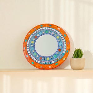 Peut inclure: Miroir décoratif rond avec un cadre orange et un motif mosaïque. Le miroir présente une surface réfléchissante centrale entourée d'éléments bleus, verts et jaunes. Une petite plante en pot est placée à droite du miroir.