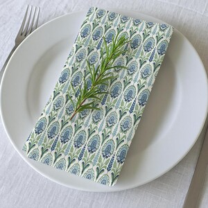 Puede incluir: Una servilleta de tela blanca con un patrón floral azul y verde. La servilleta está doblada y colocada sobre un plato blanco con una ramita de romero encima.
