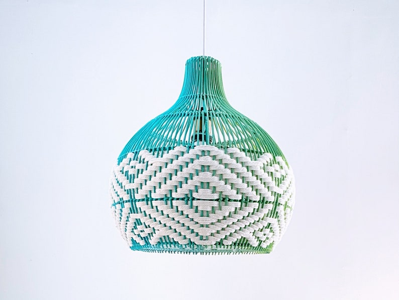 May include: A teal and white woven pendant lampshade hangs from a cord. The shade is round with a bulbous shape and features a geometric pattern in white against a teal background. The lampshade is made of a woven material, possibly rattan or wicker, and has a gradient of color from a darker teal at the top to a lighter teal at the bottom. The overall style is bohemian and modern.