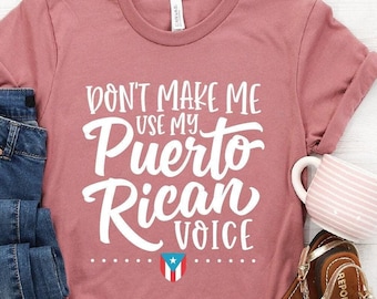May include: A pink t-shirt with the text "Don't make me use my Puerto Rican voice" in white lettering. A small Puerto Rican flag is below the text. The shirt is laid out on a white surface with a pair of blue jeans, a mug, and sandals.