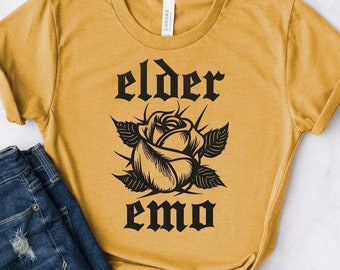 May include: Mustard yellow t-shirt with the words "elder emo" in black gothic font, with a black rose graphic. The shirt is paired with denim shorts and sunglasses.