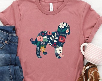 May include: A mauve t-shirt with a floral dog silhouette design. The dog is outlined in black and filled with a variety of colorful flowers. The t-shirt is paired with blue jeans and brown sandals.