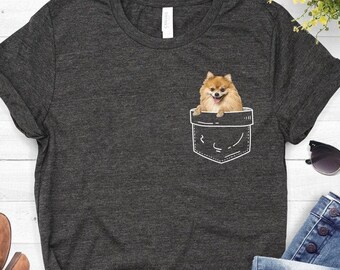 May include: A dark gray t-shirt with a white pocket design featuring a small, orange Pomeranian dog. The dog is sitting inside the pocket.
