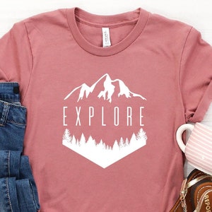 May include: A dusty rose t-shirt with rolled sleeves, featuring a white graphic of mountains and trees with the word "EXPLORE". The shirt is paired with blue jeans, brown sandals, and a pink and white striped mug.